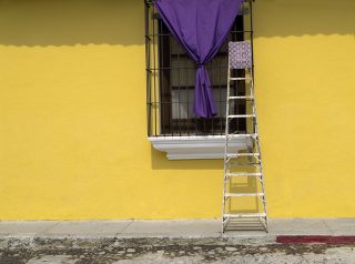 A ladder leans against a window with a purple curtain in the middle of a bright yellow wall