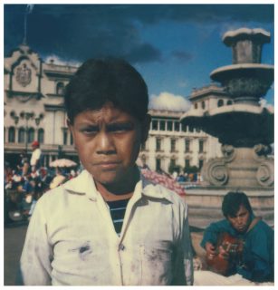 A boy with a furled brow looks disapprovingly while standing in front of a musician playing guitar on a fountain in open square